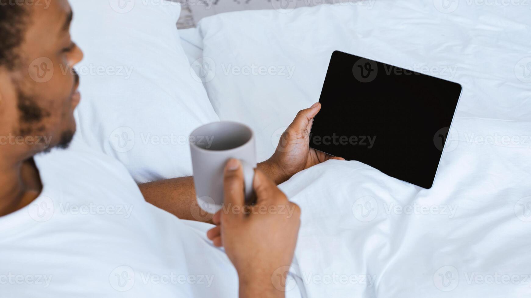 African American Man Using Digital Tablet With Empty Screen Enjoying Morning Coffee And Reading E-Book Online Lying In Bed At Home On Weekend. Internet Website Advertising. Cropped, Mockup, Panorama photo