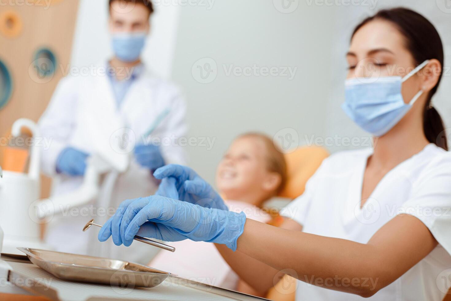 Nursing work and prepare of instruments for patient examination. Busy millennial woman in white coat, protective mask and gloves holds tool, little girl in chair and doctor on blurred background photo