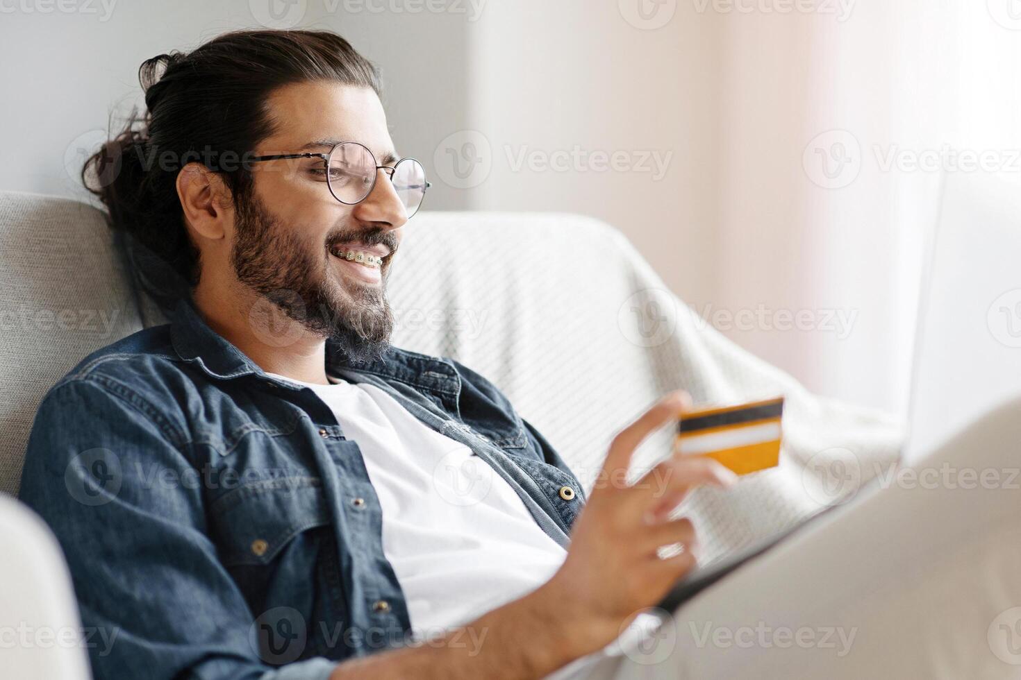 Cheerful Indian Guy In Eyeglasses Using Laptop And Credit Card At Home For Online Payments, Sitting On Comfortable Couch, Transferring Money, Shopping In Internet Or Booking Vacation, Free Space photo