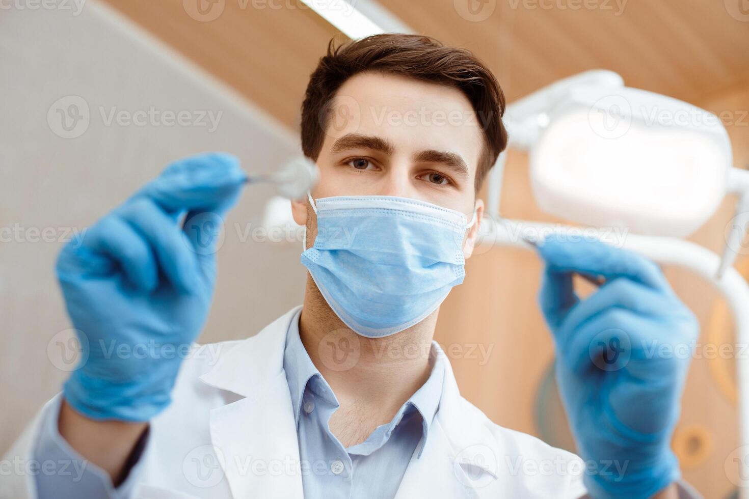 Dentistry profession, doctor treatment, curing, help skilled worker and health medical. Professional male dentist in white coat, protective mask and gloves hold dental instruments and start job photo