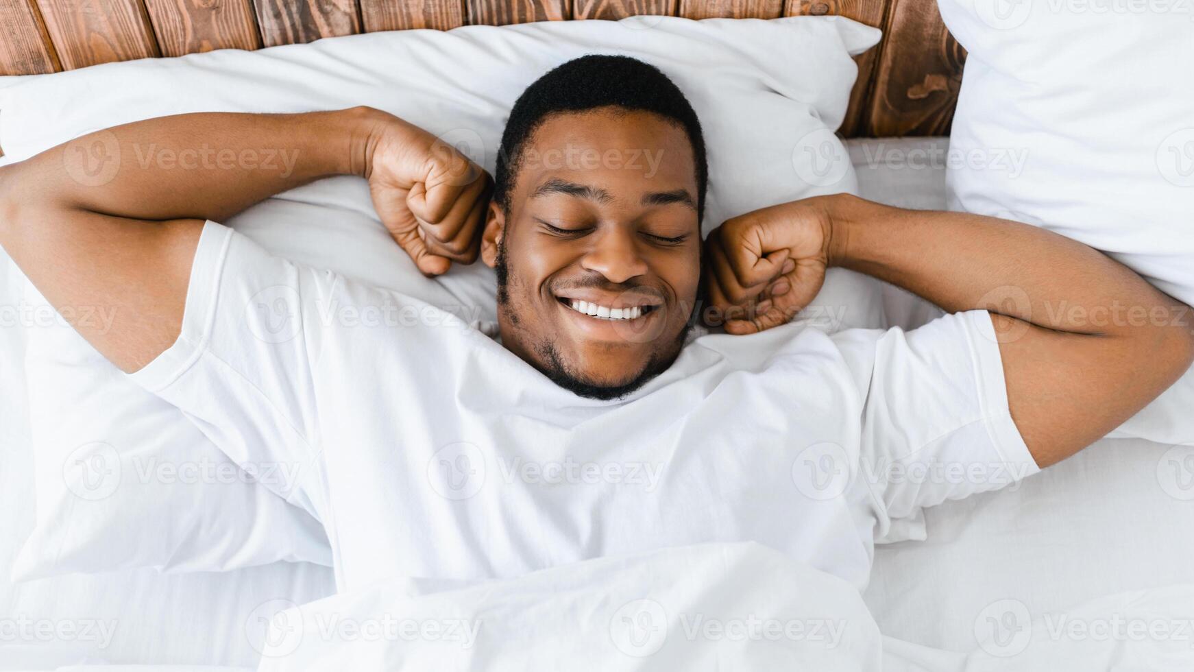Good Morning. Happy Black Man Waking Up Stretching Hands Lying In Bed At Home, Smiling With Eyes Closed. Top-View, Panorama photo