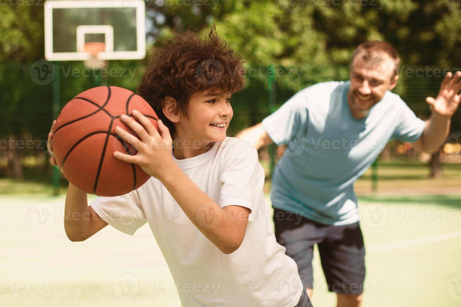Active Leisure Concept. Portrait of cheerful father and son playing basketball together, man trying to catch the ball, boy running from him, selective focus, blurred background photo