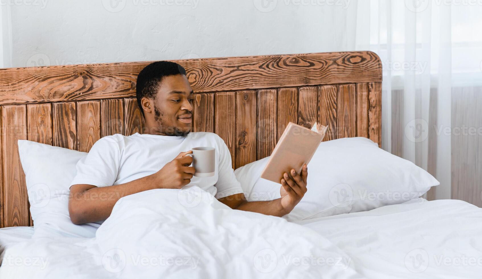 Morning Read. Black Millennial Guy Reading Book Drinking Coffee Lying In Comfortable Bed In Bedroom At Home. Weekend Relaxation And Leisure, Longread Novel Concept photo