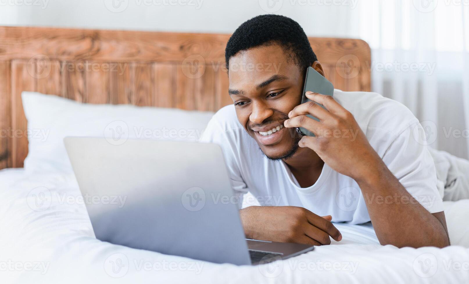 Always In Touch. Black Man Talking On Cellphone Using Laptop Computer Lying In Bed In Bedroom At Home. Phone Call, Freelance Lifestyle And Distant Communication Concept. Selective Focus photo
