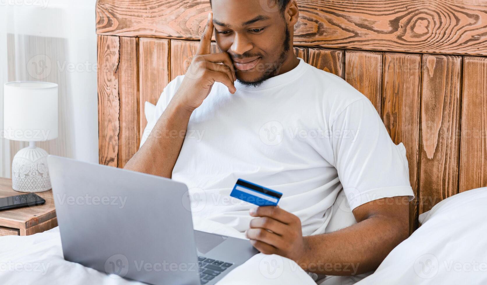 Online Shopping. Black Man With Laptop And Credit Card Making Order Sitting In Bed In Bedroom At Home. E-Commerce, Happy Customer Buying Things In Internet Shop Concept. Selective Focus photo