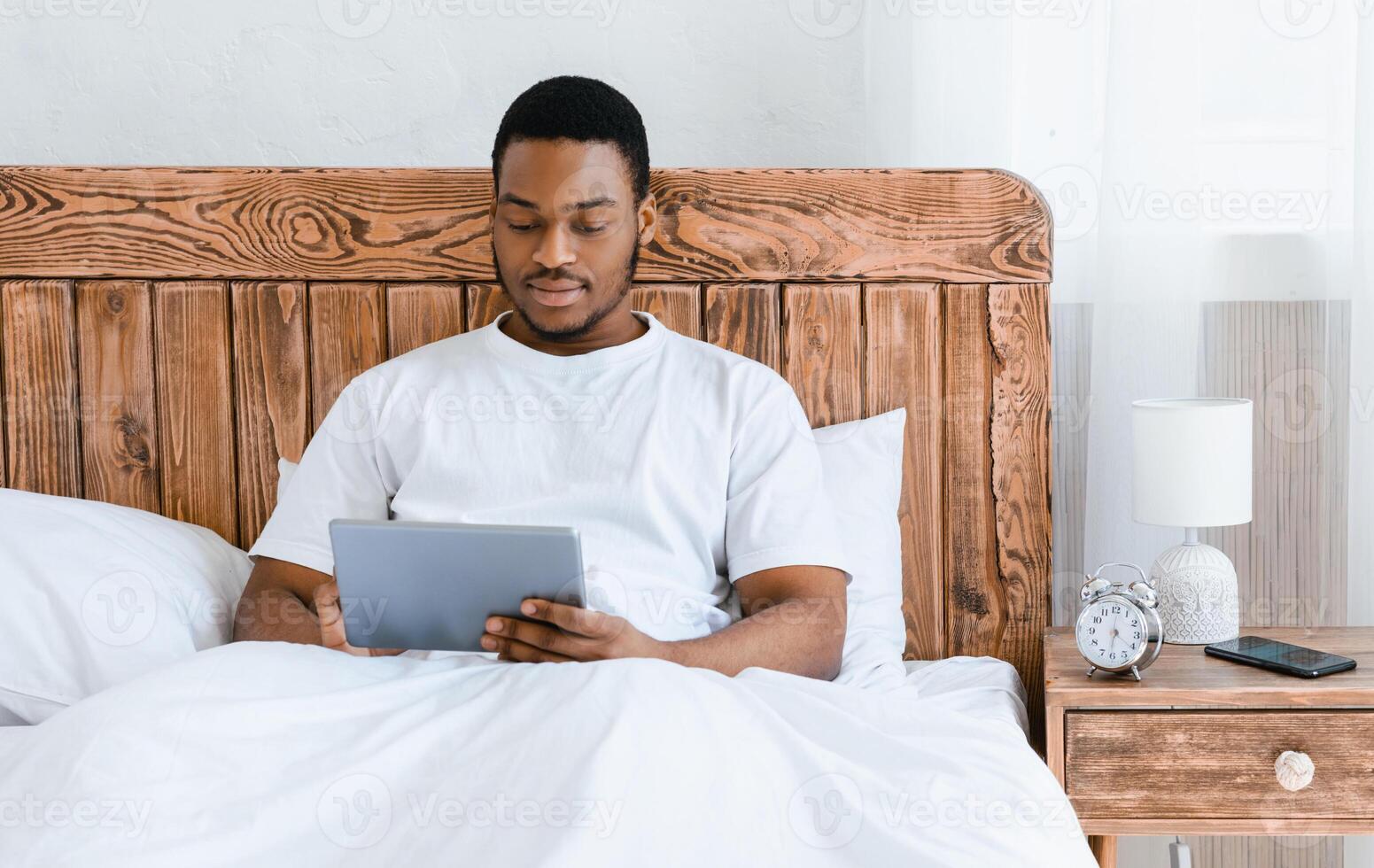African American Man Using Digital Tablet Browsing Internet Sitting In Bed In Bedroom At Home. Guy Watching Movie Online On Computer. Copy Space For Text photo
