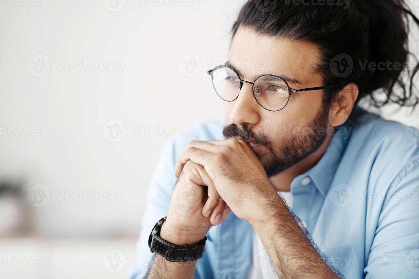 Portrait Of Pensive Millennial Indian Man In Eyeglasses Looking Away With Thoughtful Face Expression, Sitting Indoors, Young Western Guy Thinking About Business Solution, Daydreaming, Closeup Shot photo