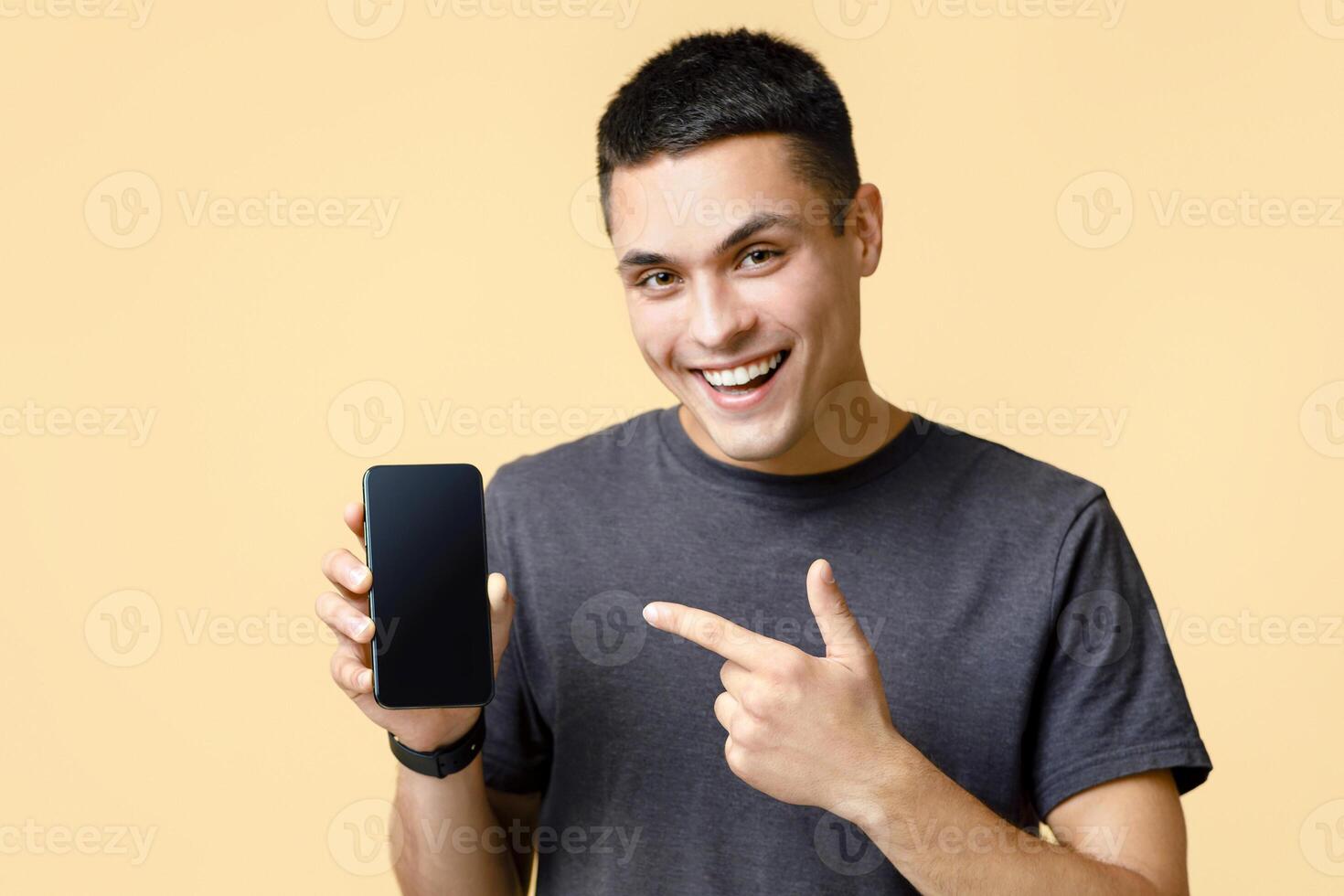 Work online, sale, modern devices and advertising of mobile application. Happy young man points finger at smartphone with empty screen, isolated on light sand background, studio shot photo