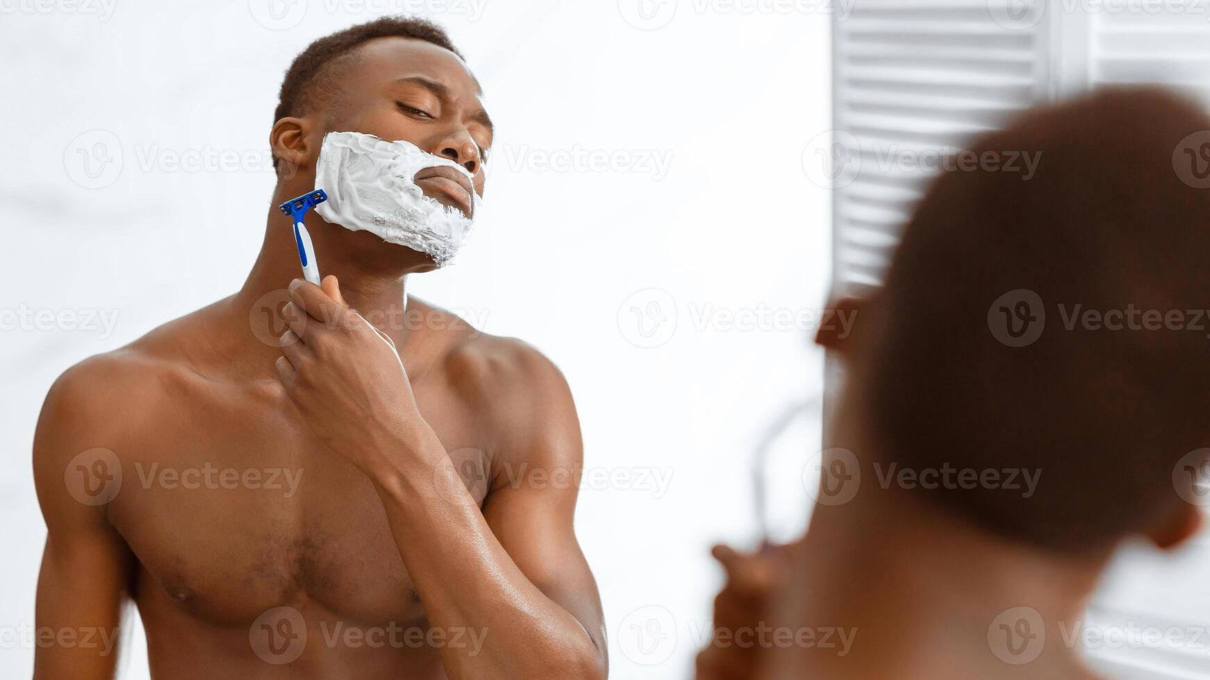 Shirtless African American Man Shaving Face With Shaver Razor And Shave Foam Standing Near Mirror In Modern Bathroom At Home. Male Facial Skincare And Morning Routine. Selective Focus, Panorama photo