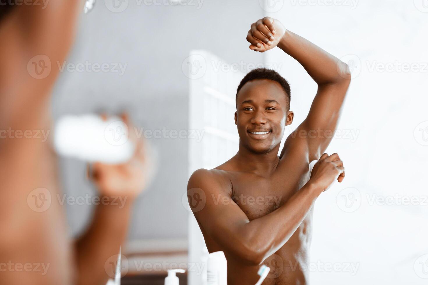 Happy Black Man Applying Deodorant Stick For Sweat And Smell Protection Standing Near Mirror In Bathroom Indoor. Male Antiperspirant, Hygiene Routine, Armpits Skincare Cosmetics. Selective Focus photo
