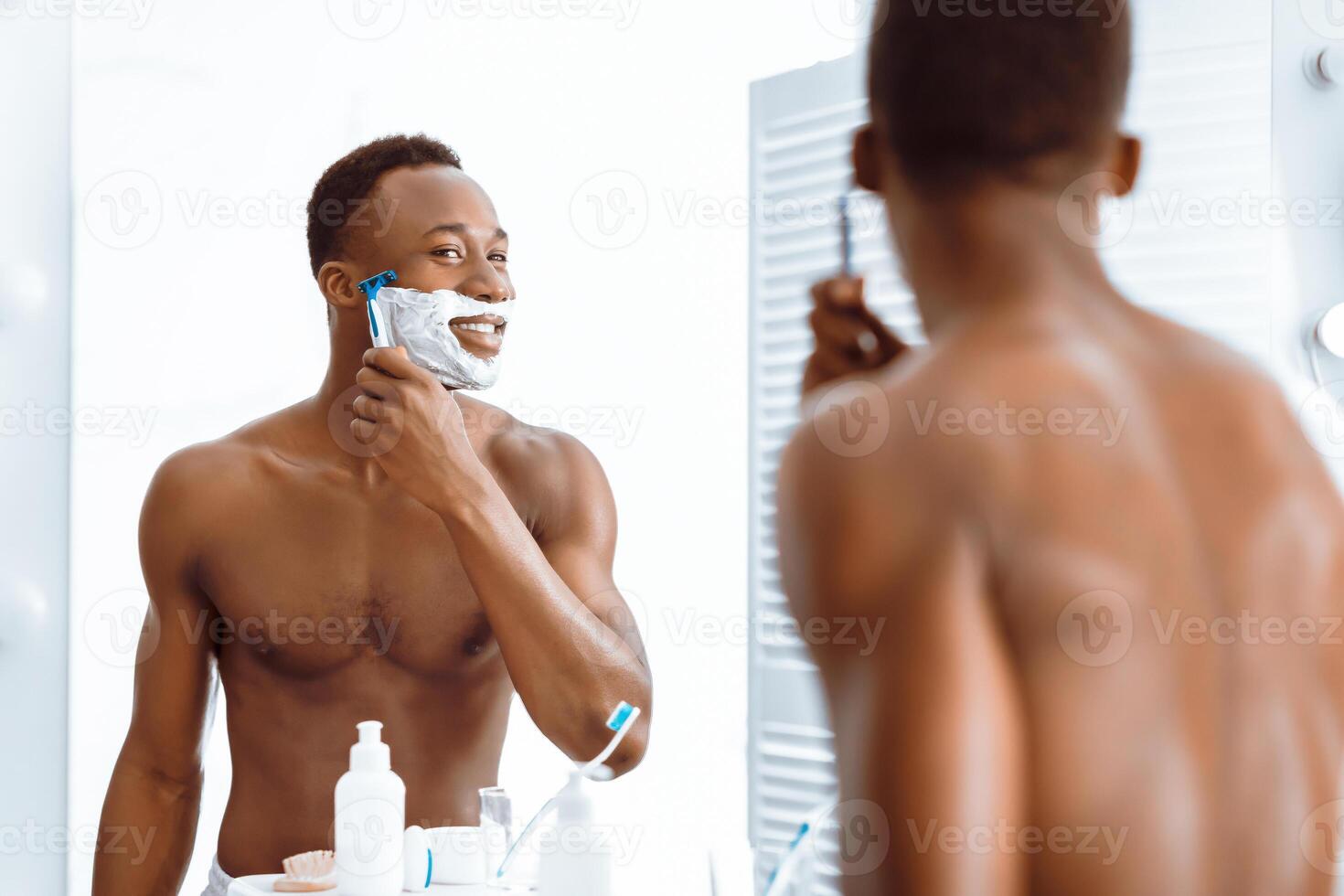 Shirtless African American Man Shaving Face Using Razor And Foam Caring For Skin, Removing Stubble Standing Near Mirror In Bathroom At Home. Male Facial Skincare And Morning Routine. Selective focus photo