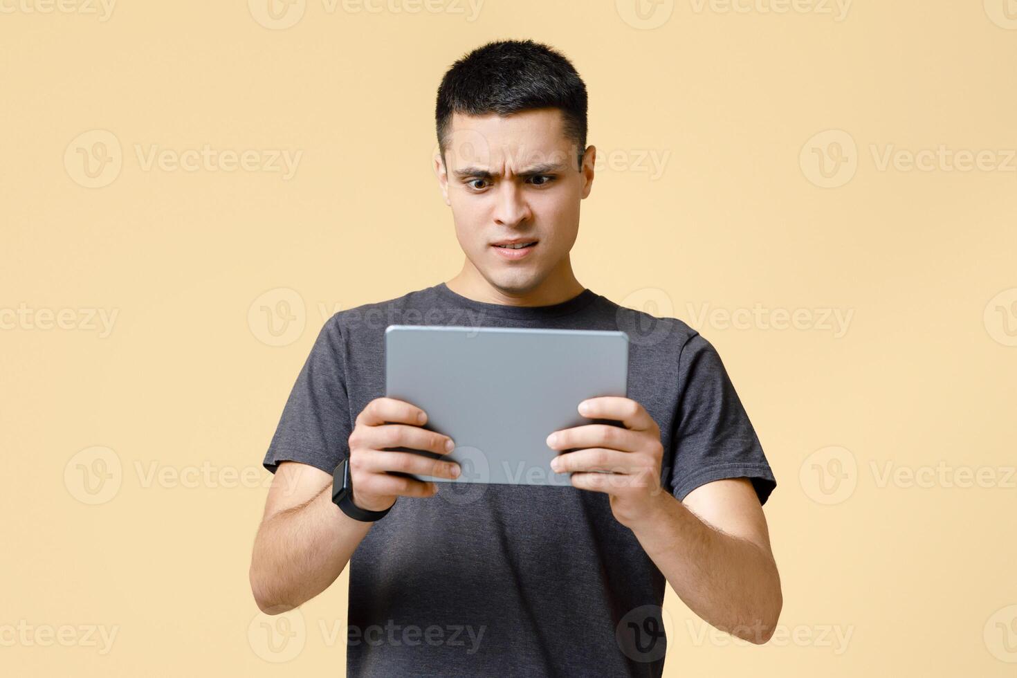 Strange , shocking information, weird message. Surprised and upset young man in brown t-shirt looks in digital tablet and reads news, isolated on beige background, studio shot, free space photo