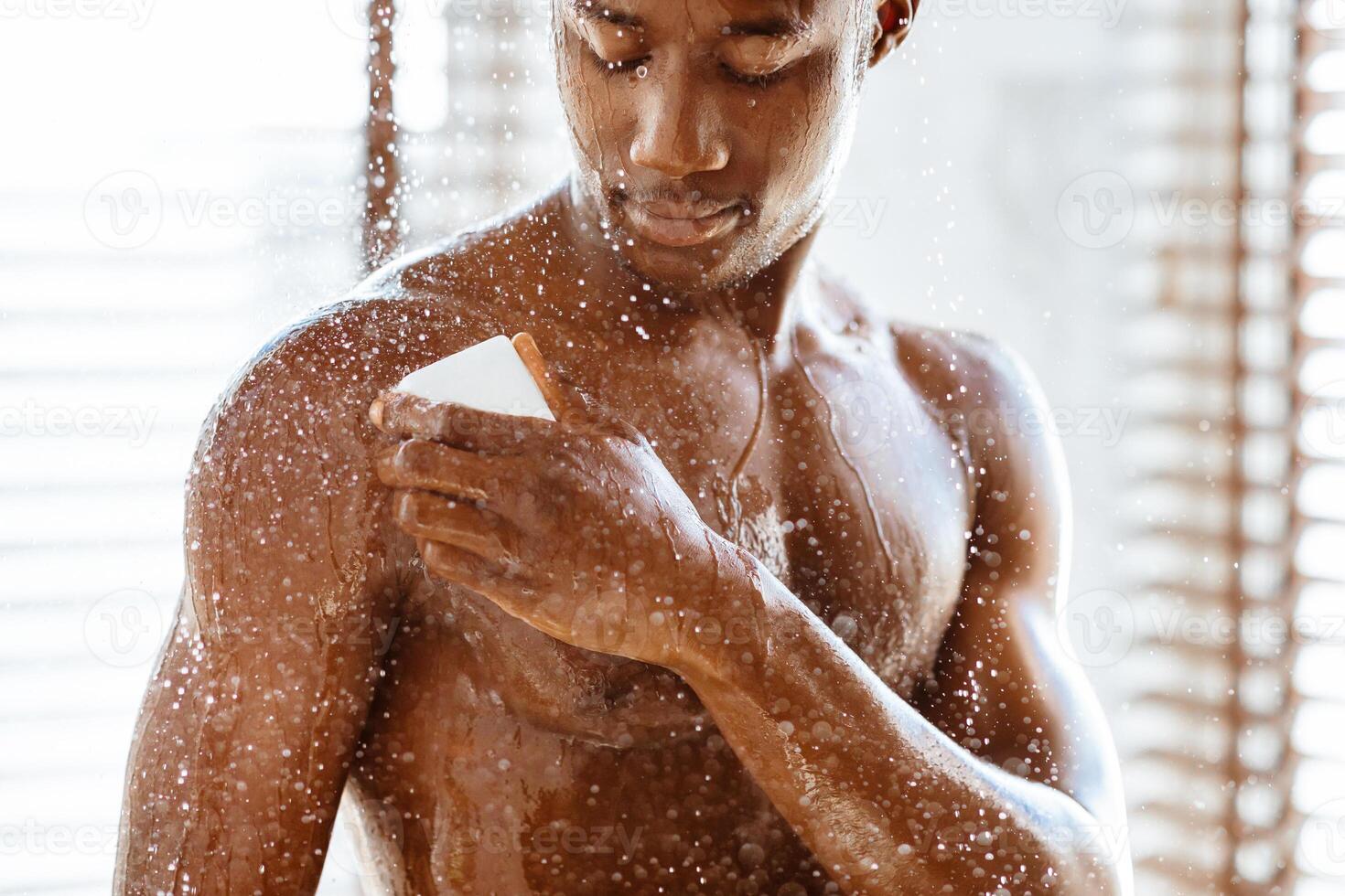 Handsome African Man Taking Shower Washing Bare Torso With Soap Standing Under Falling Water Drops In Modern Bathroom At Home. Daily Male Hygiene Routine And Bodycare Concept. Cropped photo