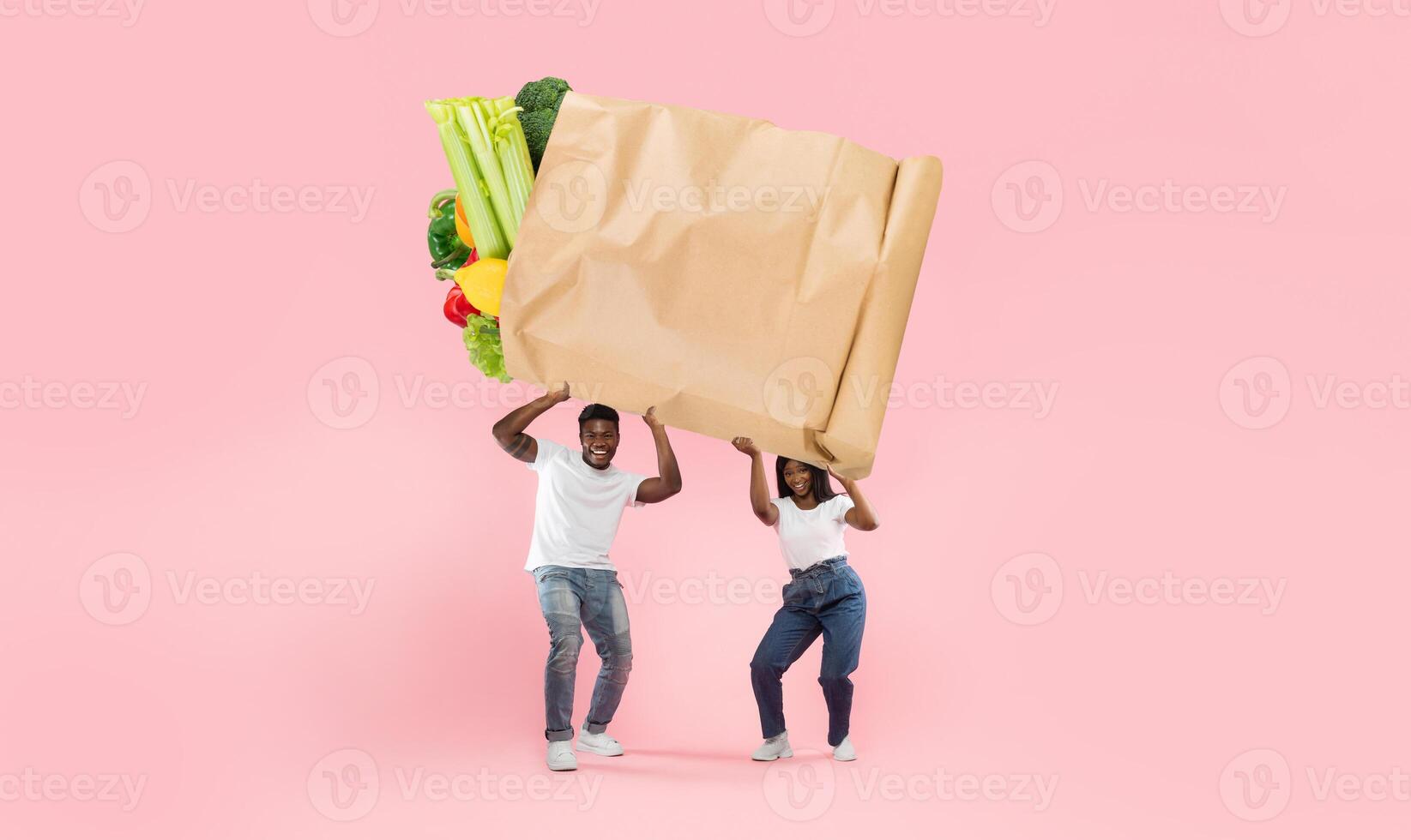 A couple energetically holds a large paper bag filled with fresh vegetables and fruits against a bright pink background. They represent the joy of grocery shopping during a new normal. photo