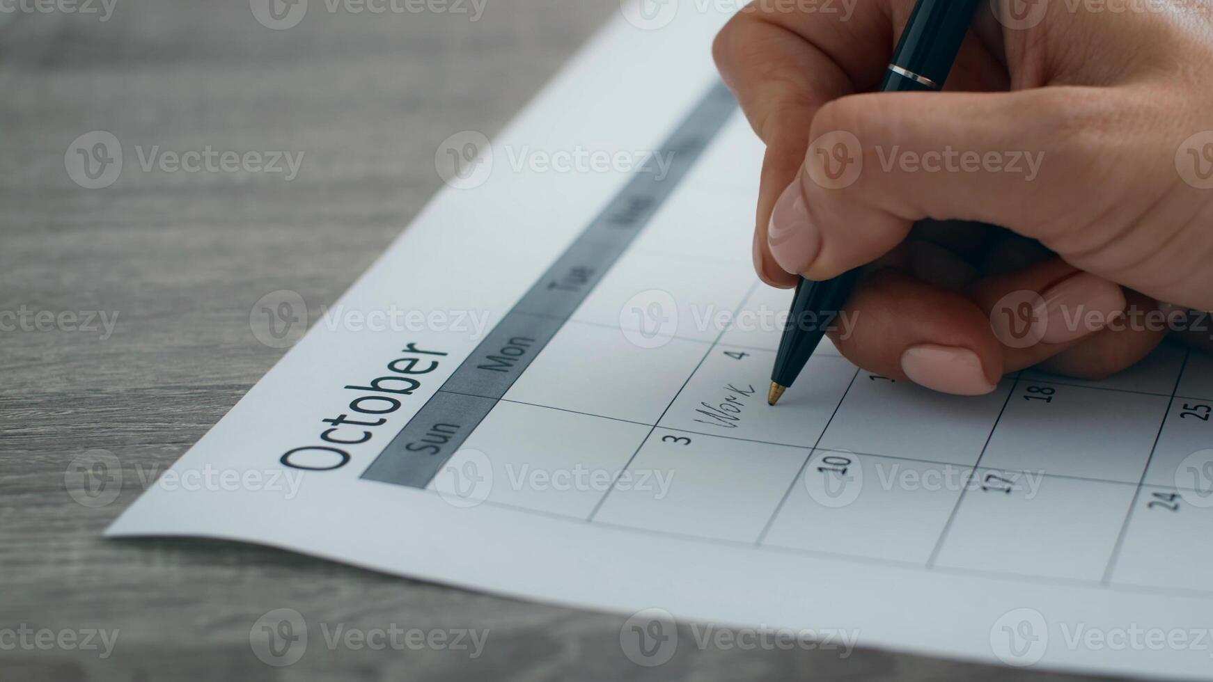 A young professional is using a pen to write on a calendar for October, organizing her tasks and appointments in a modern office setting. She focuses on her upcoming work week, ensuring productivity. photo