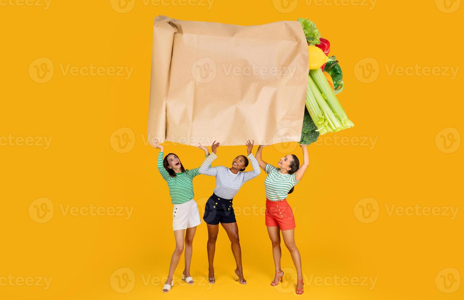 Three women joyfully raise a large paper bag overflowing with fresh groceries. They are dressed in casual clothing against a bright yellow background, reflecting the excitement of shopping. photo