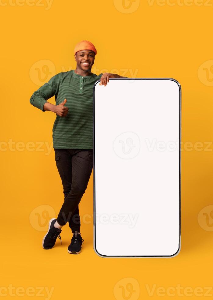 Happy Black Hipster Guy Leaning At Big Smartphone With Blank White Screen And Gesturing Thumb Up, Cheerful African Man Recommending New App Or Website, Standing On Yellow Background, Mockup Image photo