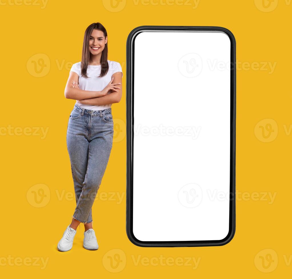 Full body length of smiling casual lady standing with folded arms near giant mobile phone, empty white screen template, orange studio background. Mock up for app or website, free space for advertising photo