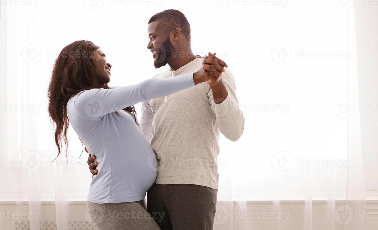 Joyful pregnant couple having fun, dancing next to window at home, free space photo