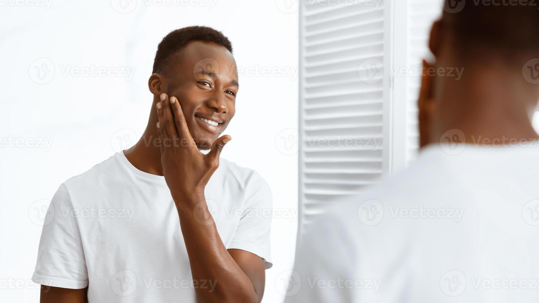 African Guy Touching Shaved Face After Shaving Looking In Mirror Standing In Modern Bathroom At Home. Male Morning Routine, Facial Skincare Cosmetics Concept. Panorama, Selective focus photo