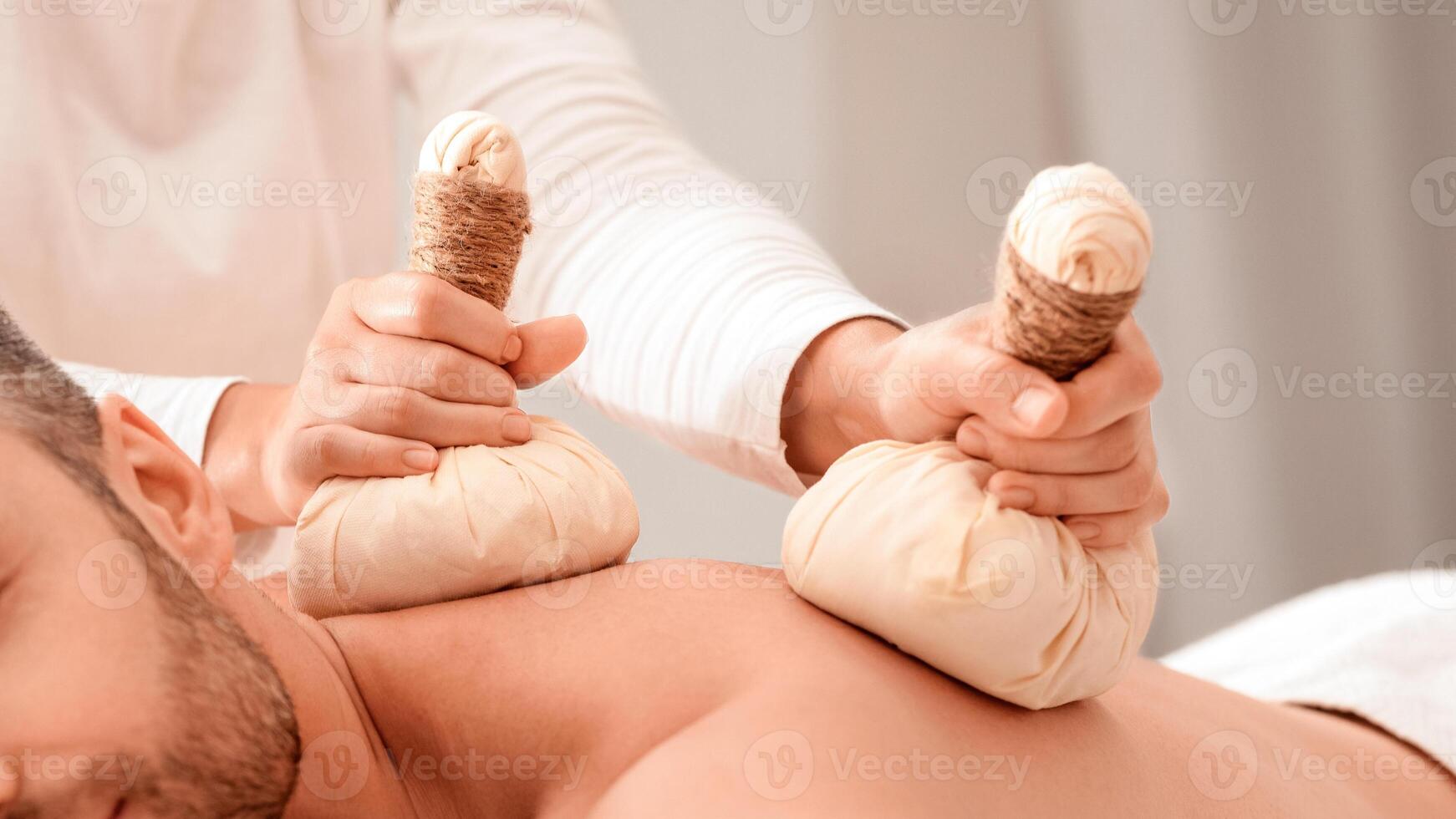 Herbal massage concept. Unrecognizable spa attendant making body massage for handsome man with herbal compress balls, luxury spa interior. Man enjoying herbal treatment at massage salon photo