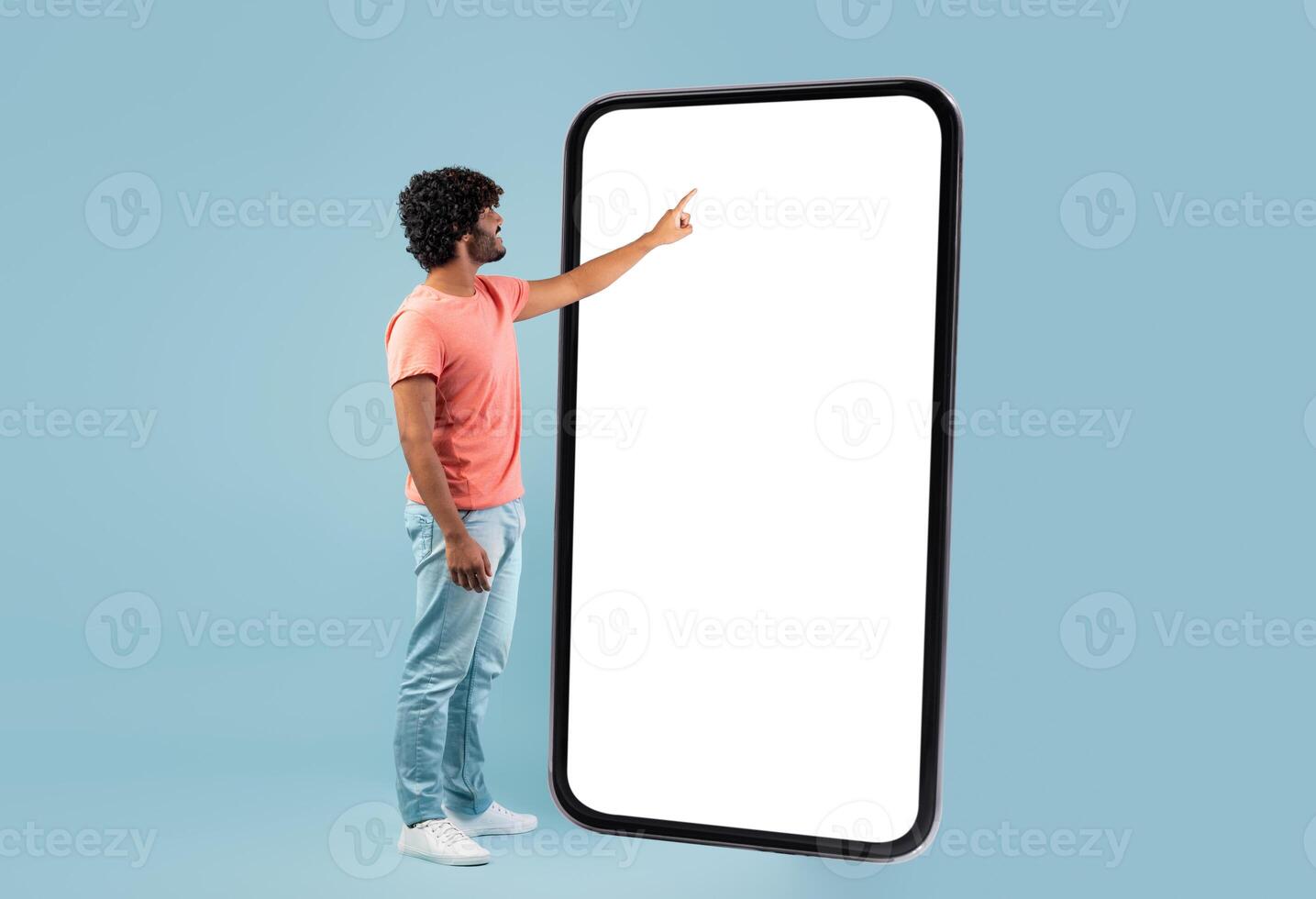 Stylish curly young hindu guy in casual outift touching huge smartphone white empty screen over blue studio background, checking newest mobile app, panorama with copy space, mockup photo