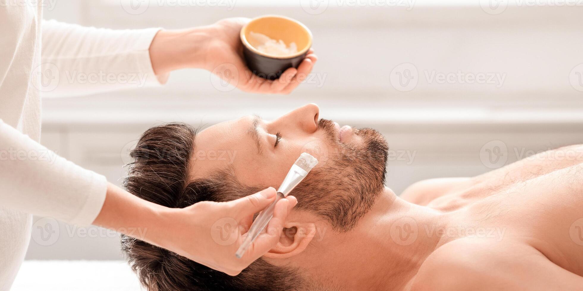 Male cosmetology and face care concept. Female attendant applying nourishing face mask on sleeping middle aged man face, spa salon interior. Side view of bearded man getting face treatment photo