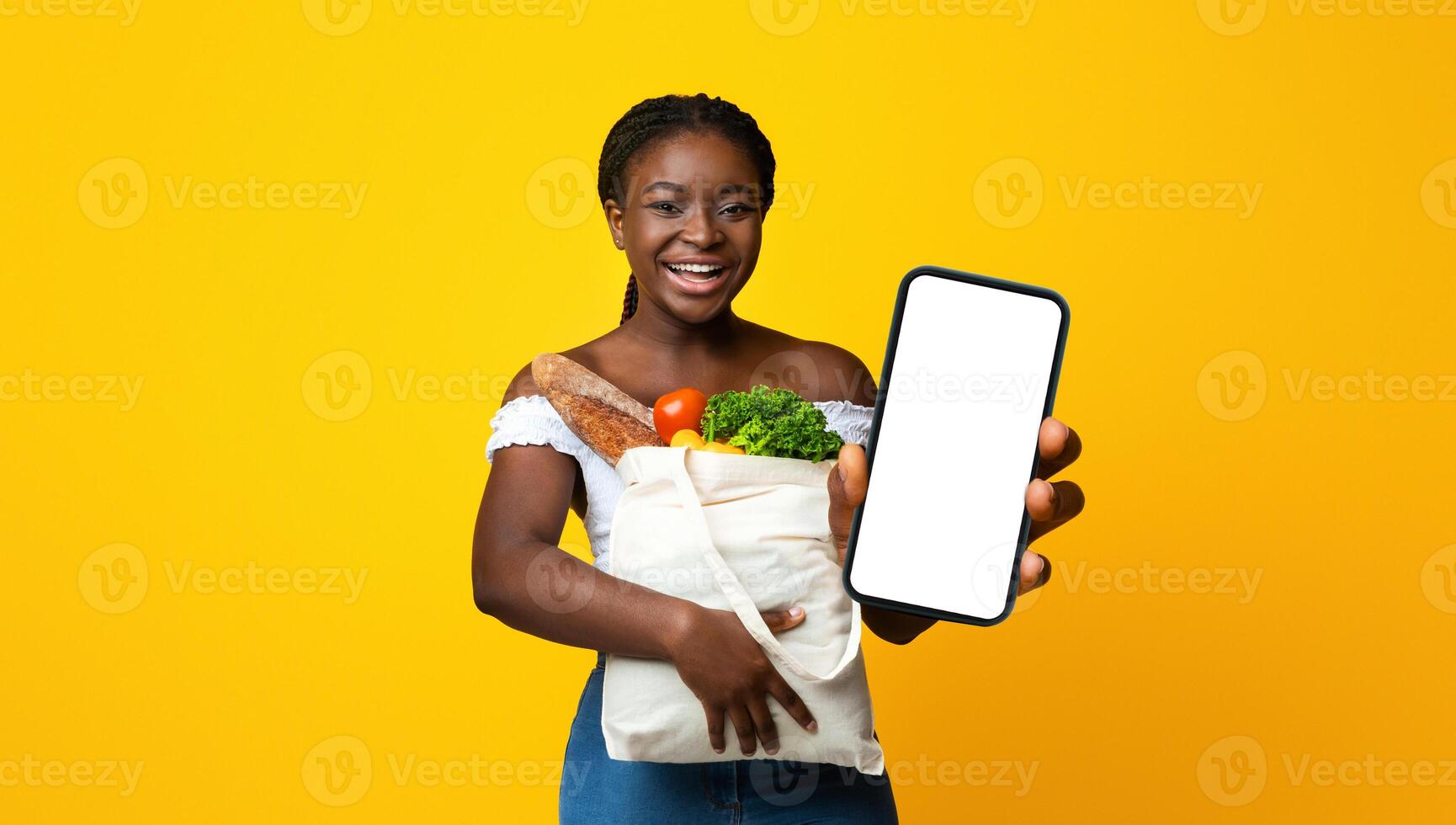 Young smiling african american woman holding paper bag full of groceries and showing thumb up, happy after successful food shopping, standing on yellow studio background, looking at camera, copy space photo