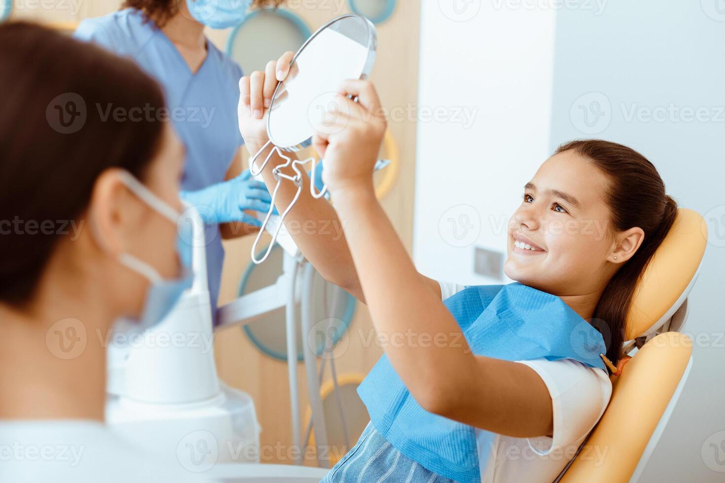 Good job and healthy teeth. Happy girl in dental chair with napkin looks in mirror in interior office with doctors in protective masks and uniforms looks at patient after treatment, free space photo