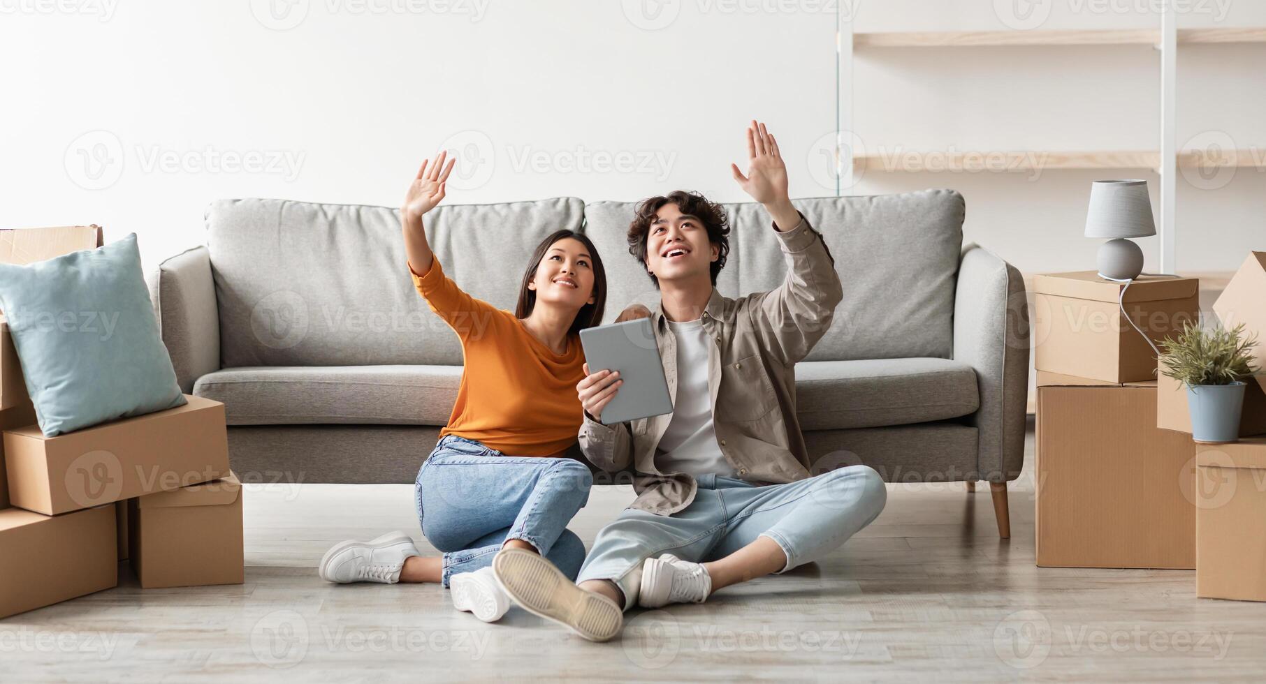 Millennial Asian couple sitting on floor among carton boxes, using digital tablet, dreaming about design of new home, imagining future furniture arrangement, panorama. Relocation, moving day concept photo