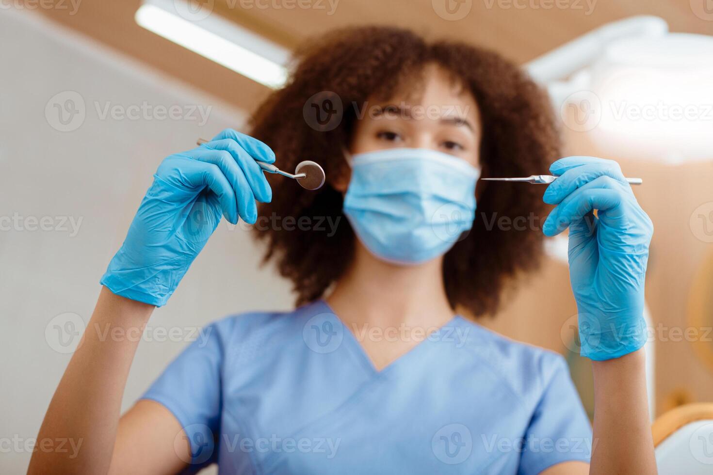 Portrait of young dentist in modern clinic. African american female in uniform, protective mask and gloves holds medical instruments for examination of patient, bottom view, close up, free space photo