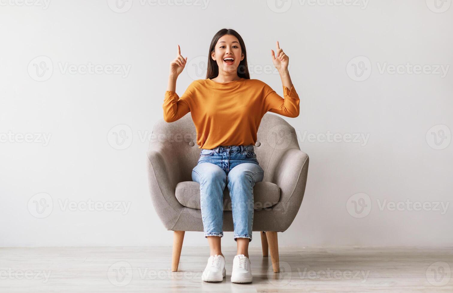 Excited young Asian woman sitting in armchair, pointing two fingers up, offering space for your advertisement against white studio wall, full length. Best promotion, cool discount, huge sale concept photo