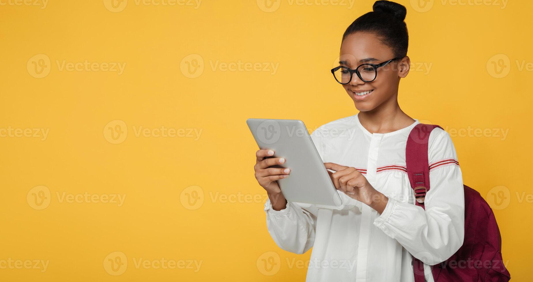 Cheerful smart teen afro american girl pupil in glasses with backpack studying with digital tablet, prepare to exam, isolated on yellow background, panorama, free space. Remote lesson, education photo