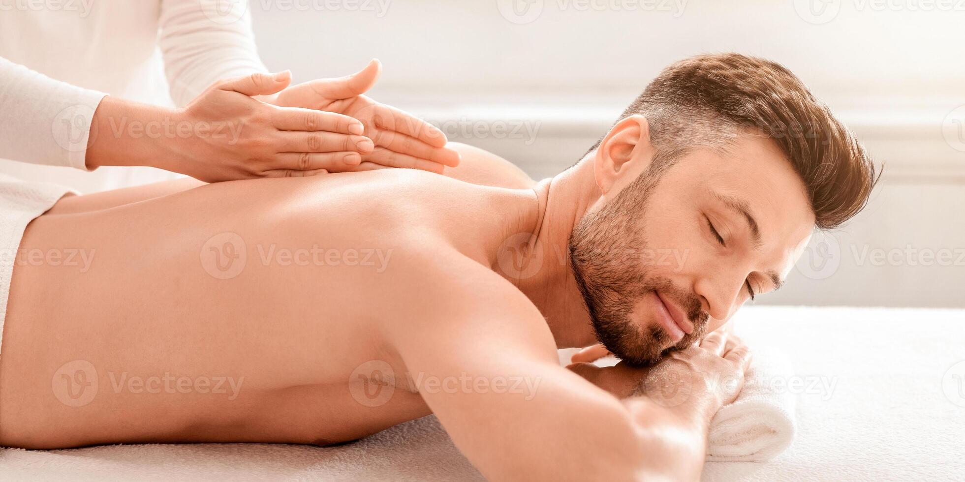 Rich middle aged man enjoying healing massage at modern spa, side view. Relaxed bearded man attending spa salon, masseuse rubbing his back, closeup. Male spa, massage salon concept photo