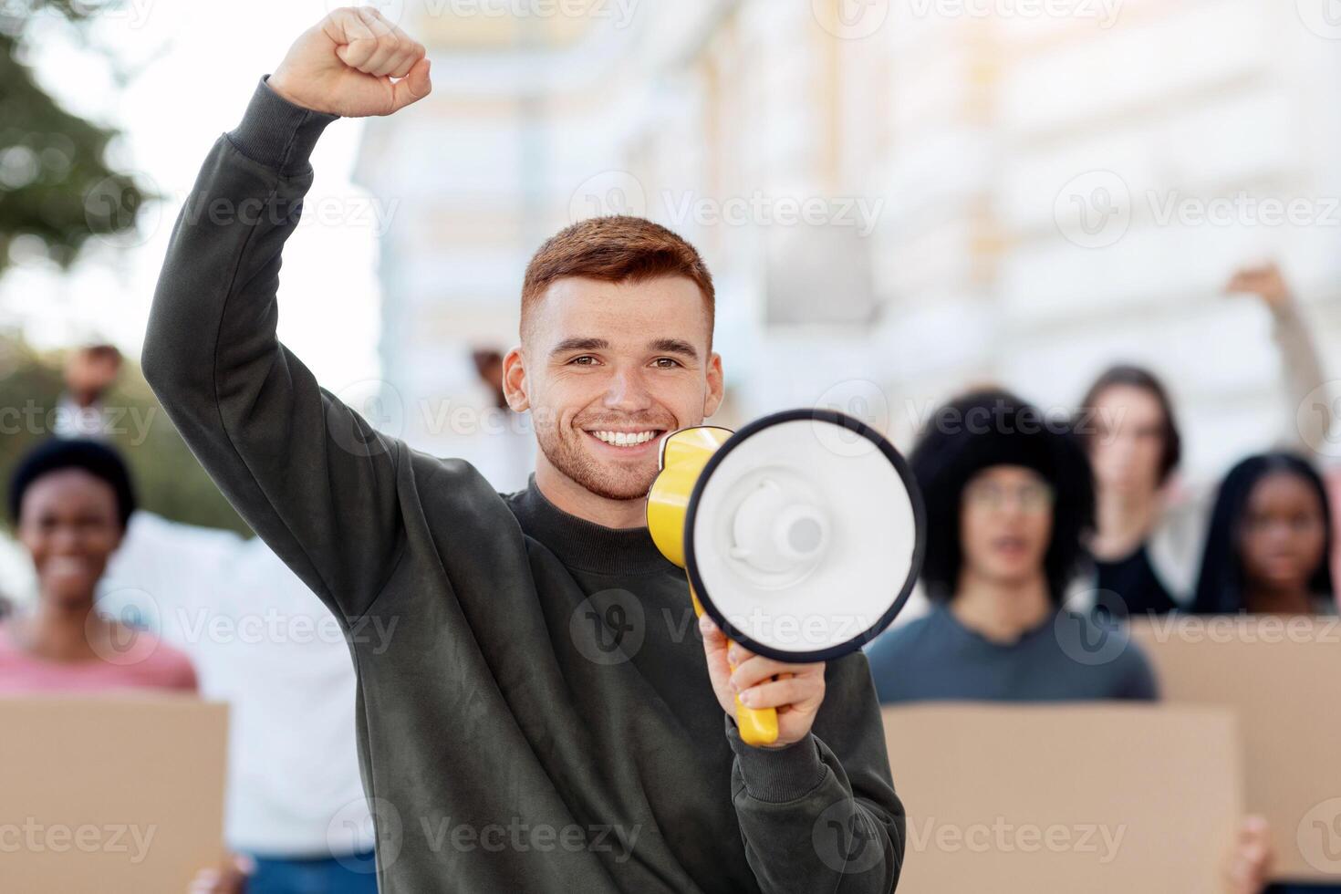 Emotional guy activist with megaphone on the street, chanting slogans and raising fist up, young caucasian man leading international group of activists, striking against lockdown rules photo