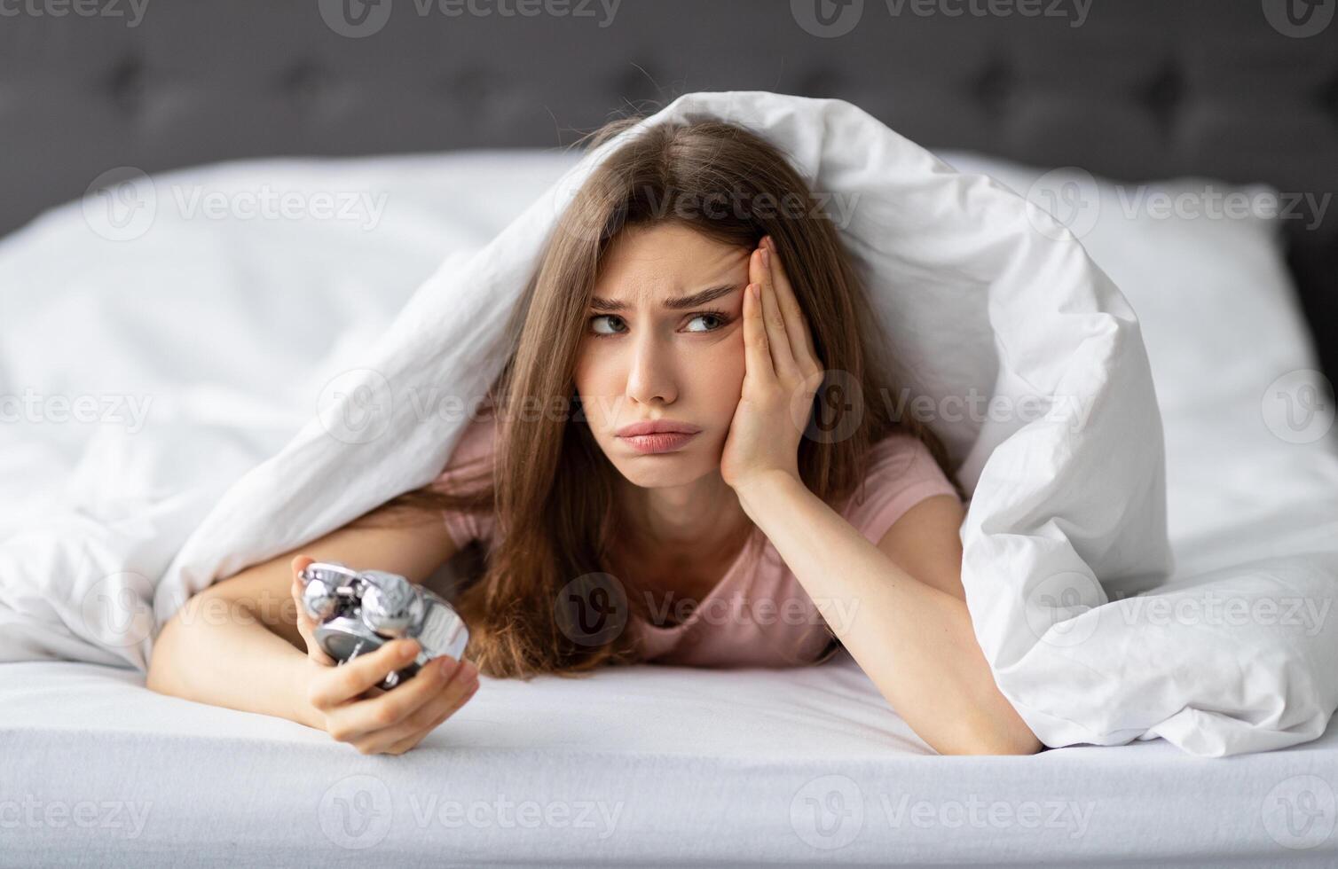 Unhappy young Caucasian woman lying in bed with alarm clock, not willing to get up in morning. Upset millennial lady having problem waking up after sleepless night, cuddling under warm blanket photo