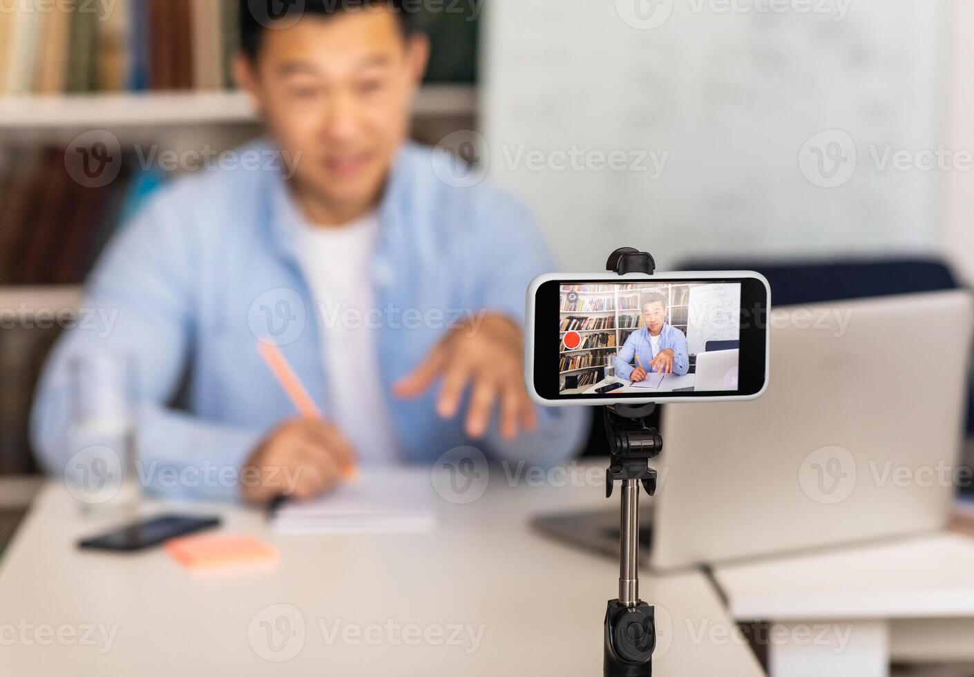 Educational Blogging. Asian Tutor Man Filming Online Lecture Via Smartphone Sitting At Workplace Indoor. E-Teaching Concept. Selective Focus On Mobile Phone photo