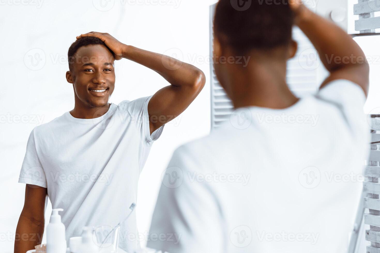 Attractive African American Guy Touching Hair Looking In Mirror After Haircare Beauty Routine Standing In Modern Bathroom At Home In The Morning. Daily Male Hair Care Concept. Selective Focus photo