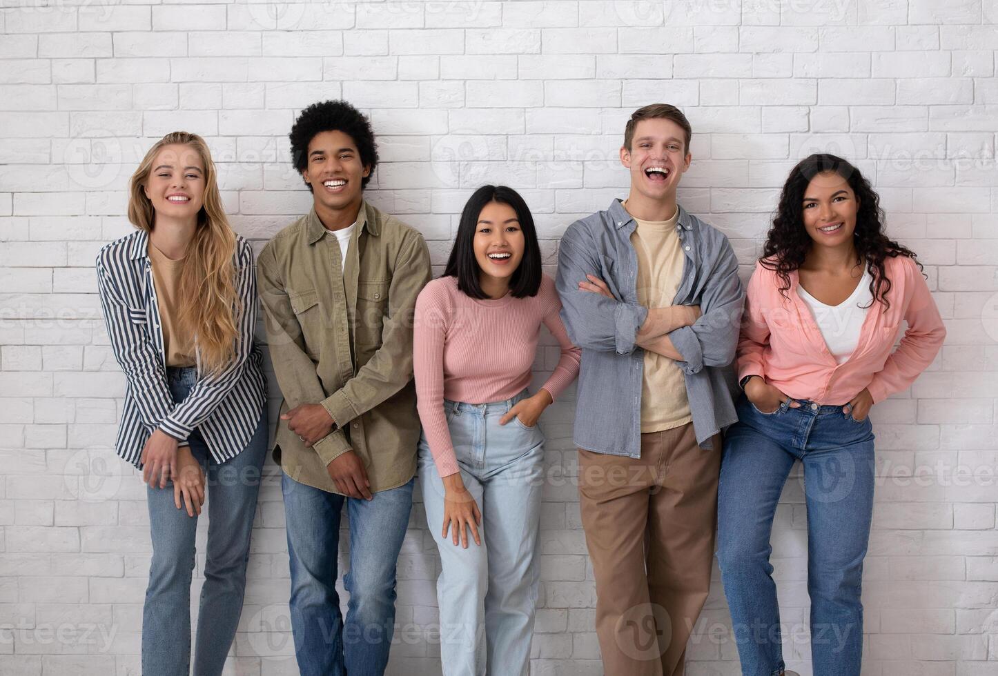 Blog about student life, friendship and modern study exchange. Cheerful teen multiracial students team in casual have fun and looking at camera, on white brick wall background, studio shot photo