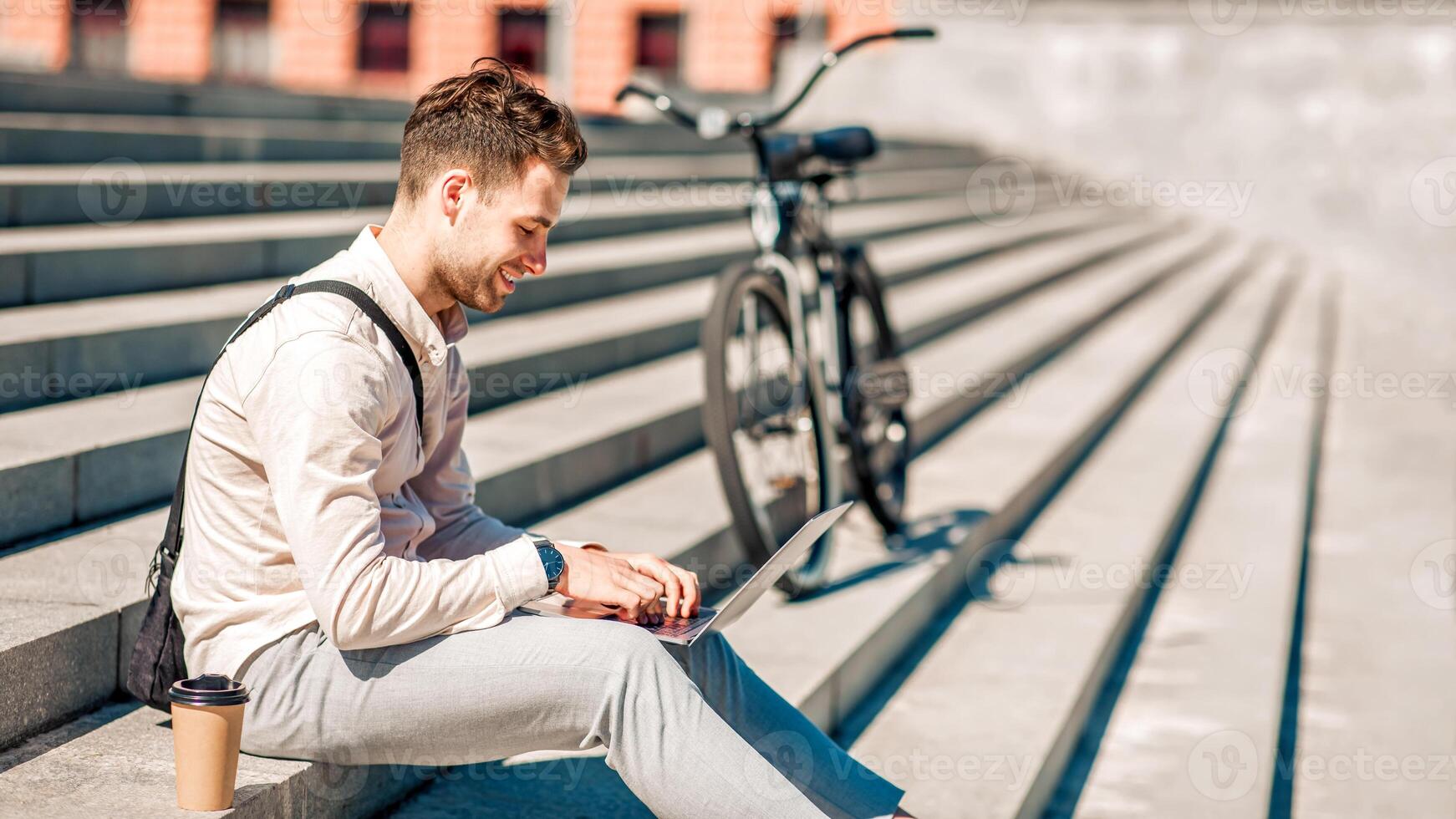 Modern employee sitting on stairs and working outdoors with takeaway coffee and bike, side view, copy space photo