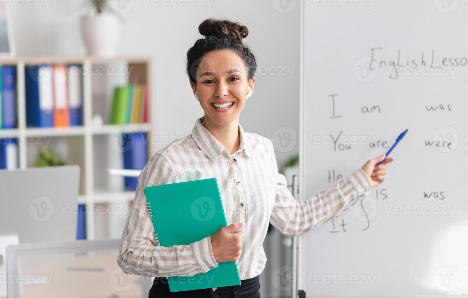 Training concept. Happy female tutor teaching English language, pointing at grammar rules on board and smiling at camera, holding clipboard in office interior photo