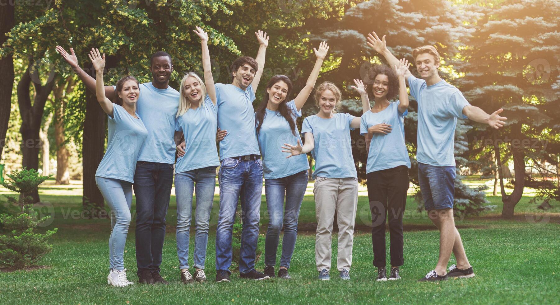 Happy millennial volunteers greeting you, waving hands and cuddling together at park, free space photo