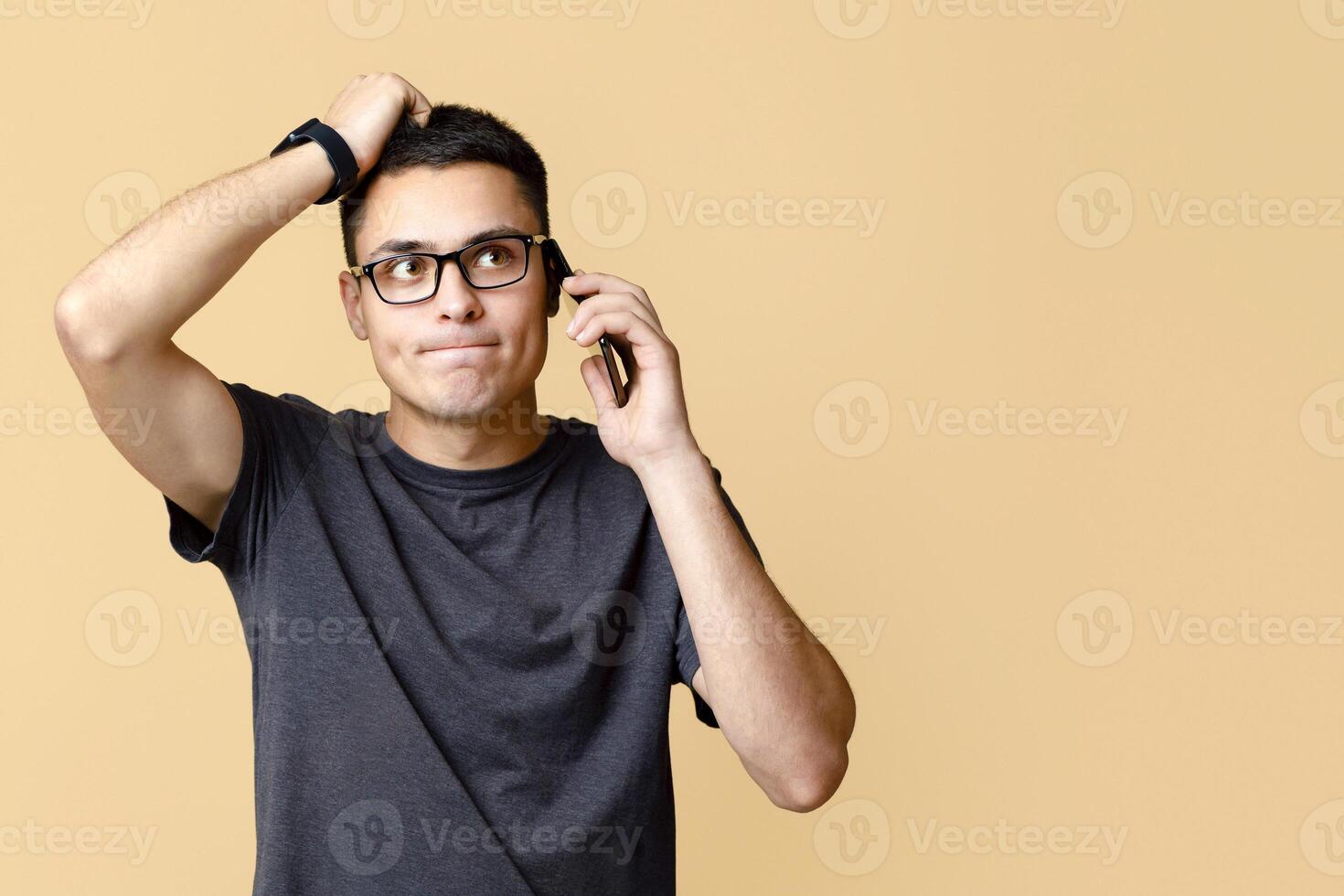 Strange news, problems and difficulties at work. Pensive sad young guy in brown t-shirt and glasses, talking on phone, holding his head and looking up at empty space, isolated on beige background photo