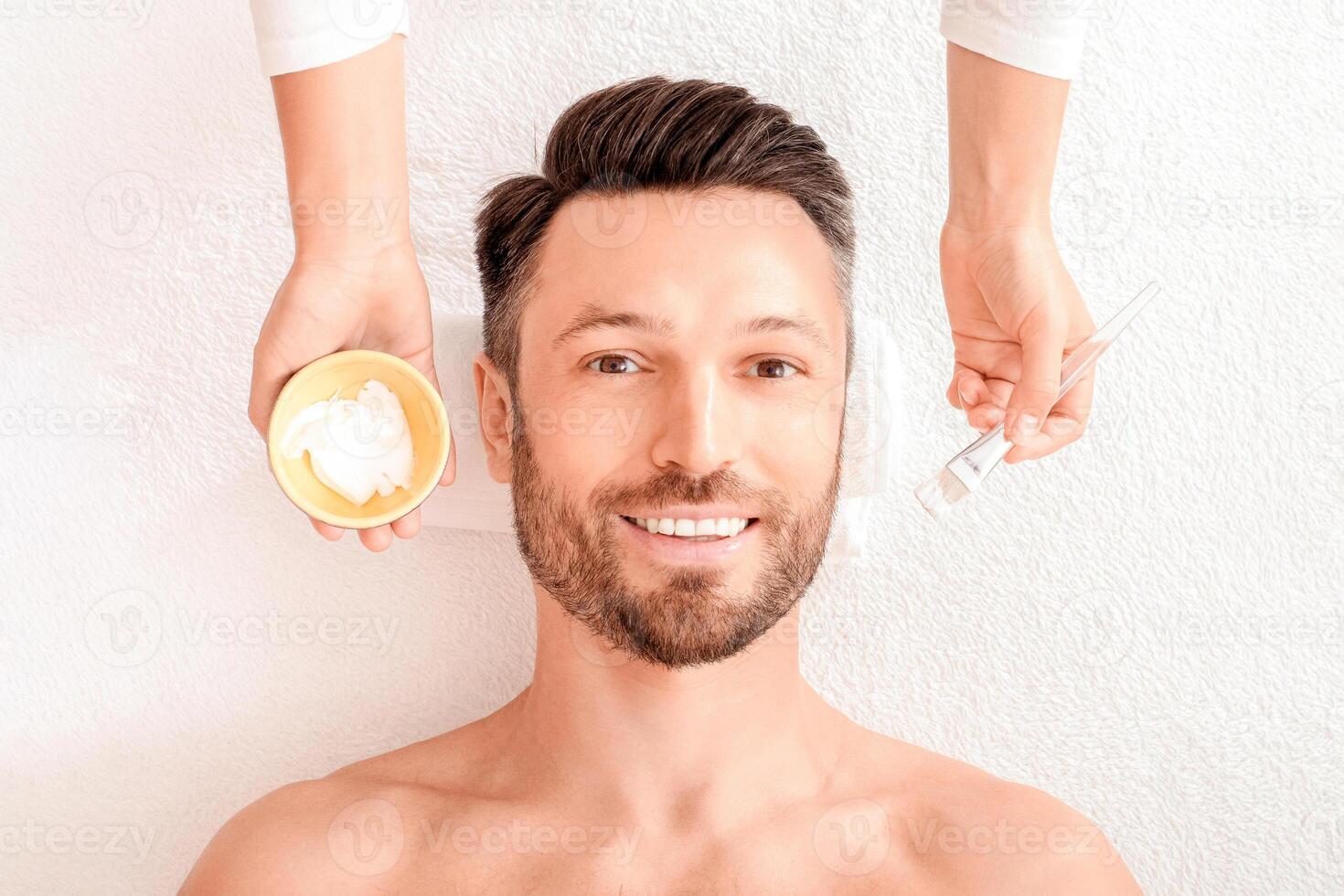Top view of bearded cheerful man getting face treatment at modern spa salon. Female attendant applying white face mask on middle aged man face. Modern male face care concept, copy space photo