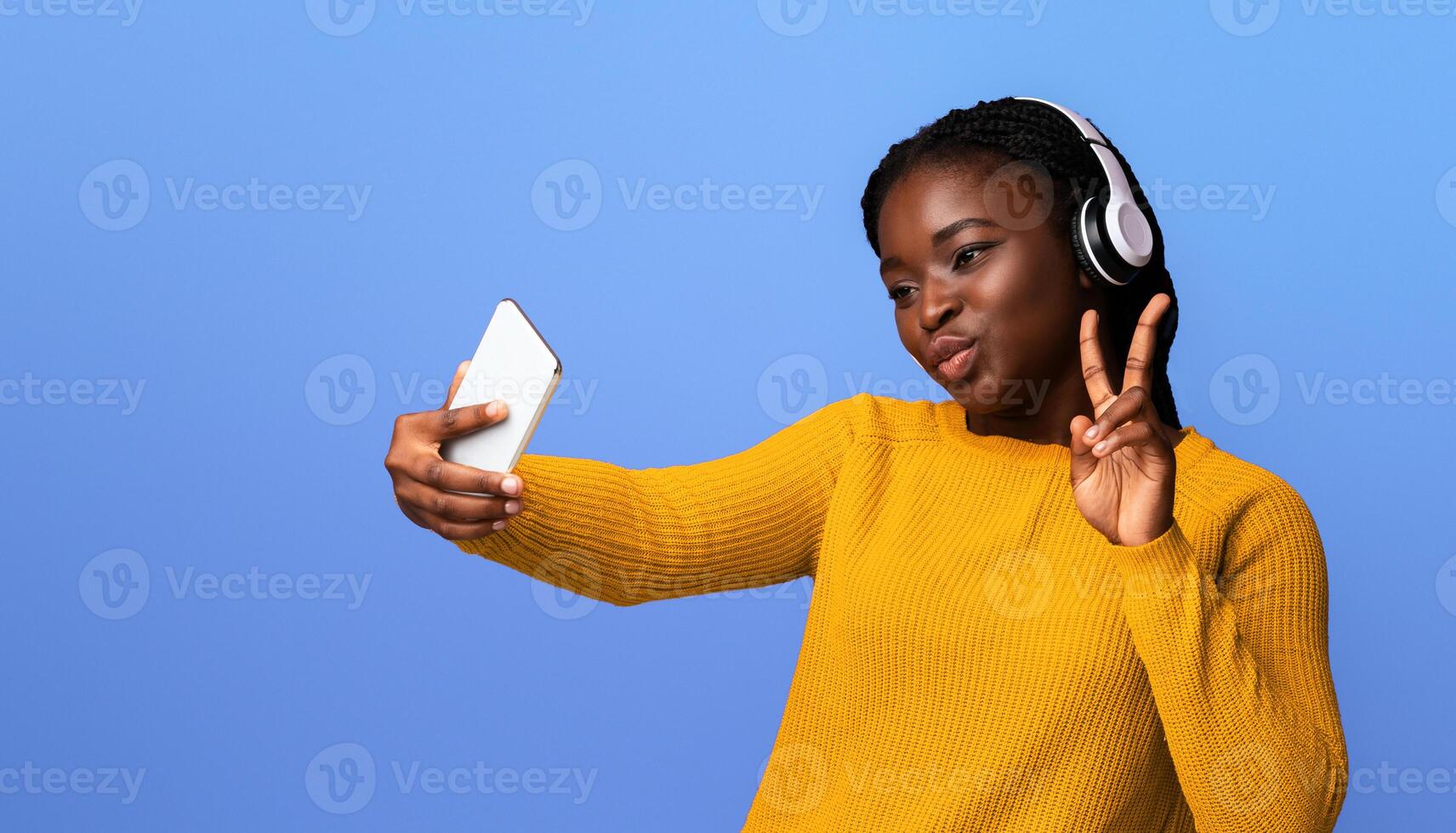 Cheerful Black Woman In Headphones Taking Selfie On Smartphone And Showing Peace Gesture While Posing At Camera, Joyful African Lady Having Fun, Posing Over Purple Background In Studio, Copy Space photo