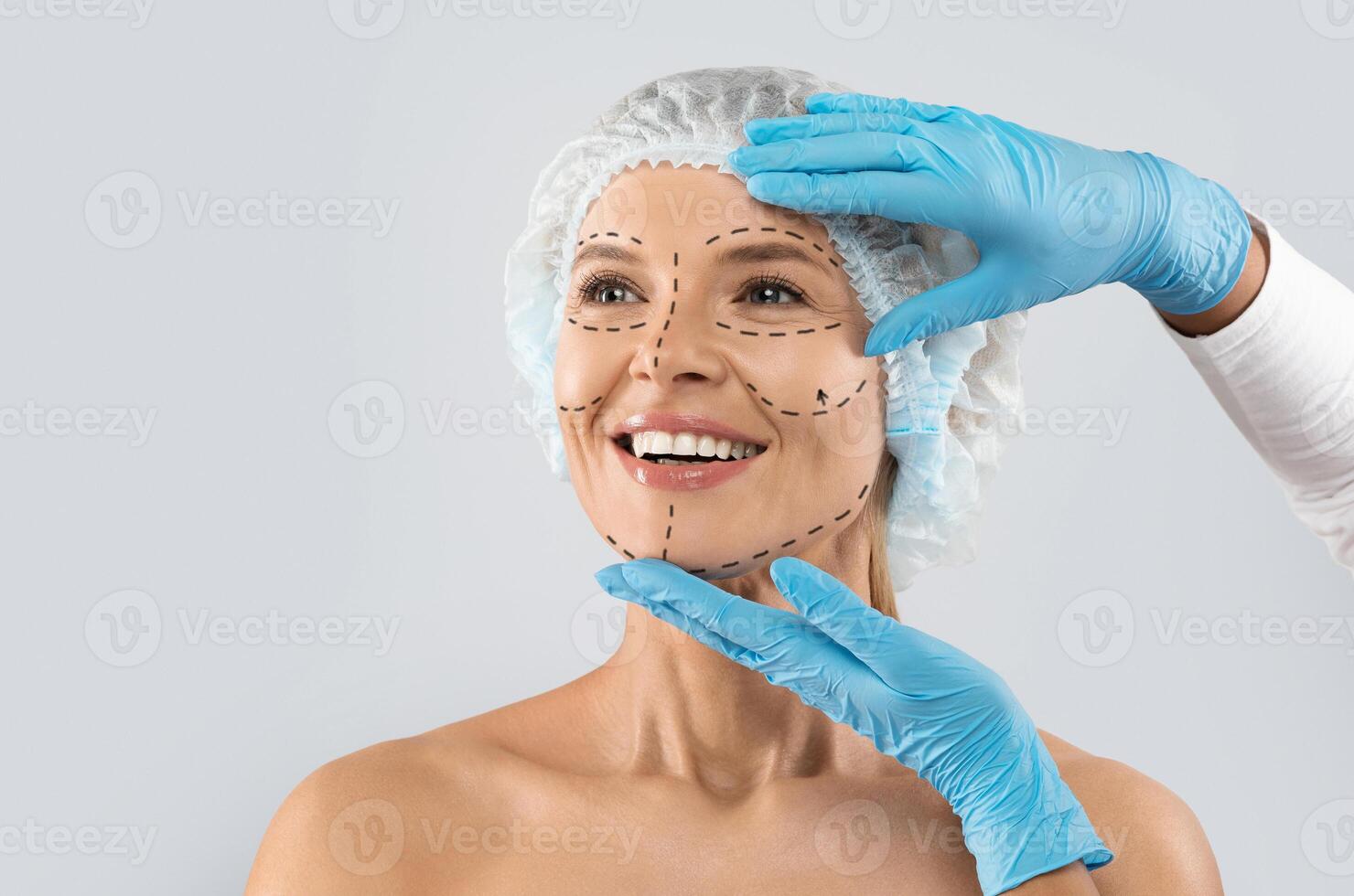 Cheerful excited middle aged woman in medical hat with pre surgery marks on her face looking at copy space for ad and smiling, surgeon hands in blue gloves touching female skin photo