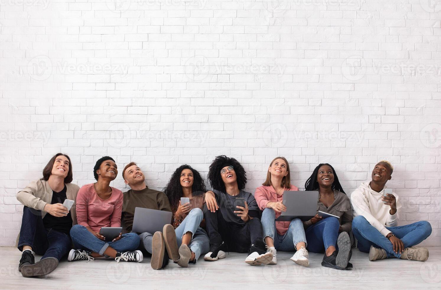 International group of friends with smartphones, laptops and digital tablets looking up on copy space, sitting on floor over white wall. Cheerful multiracial students using gadgets, studio background photo