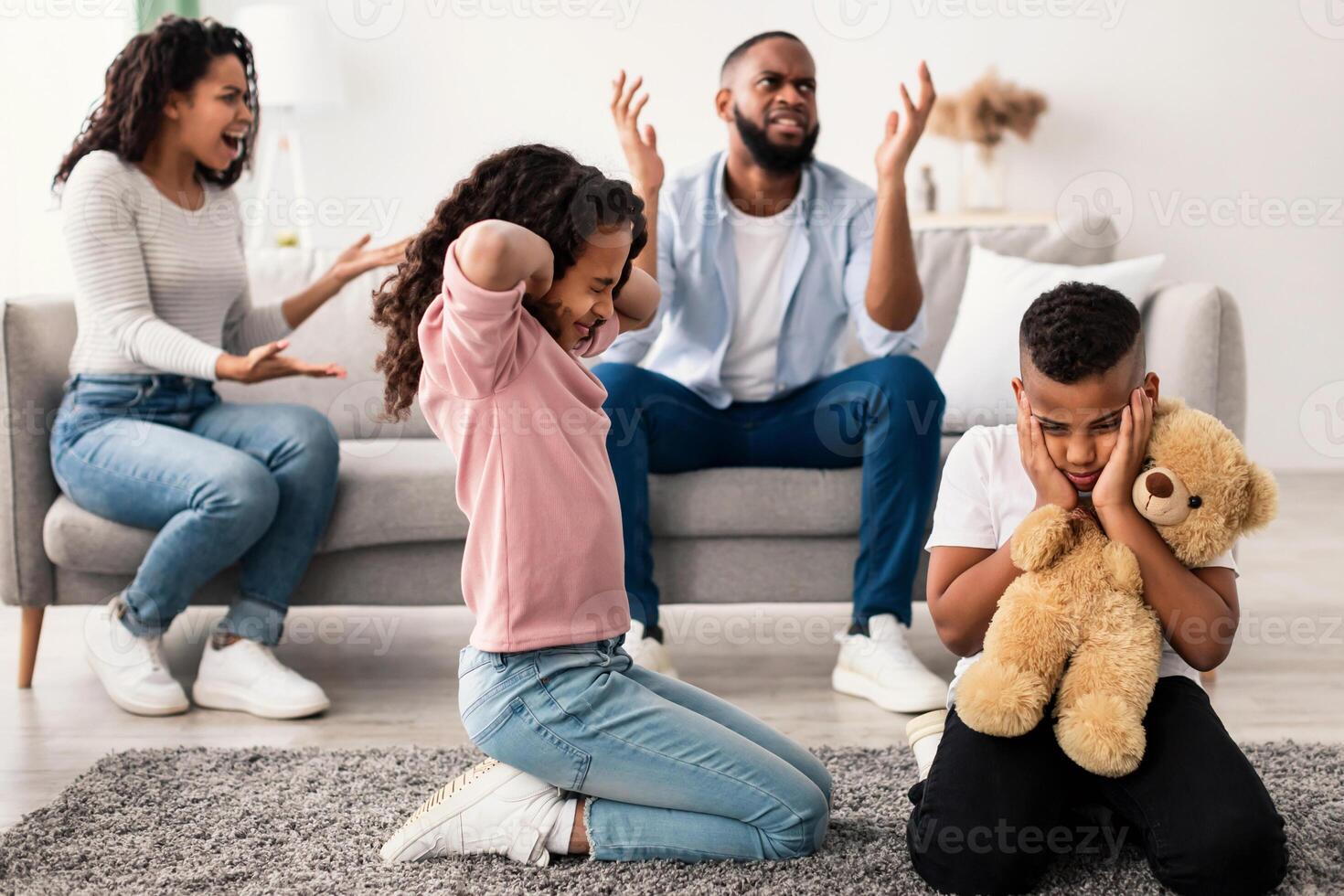 Family Conflicts. Sad little black children covering ears with hands while their parents arguing in the background, upset boy and girl don't want to hear quarrel, stressed kid sitting on the floor photo