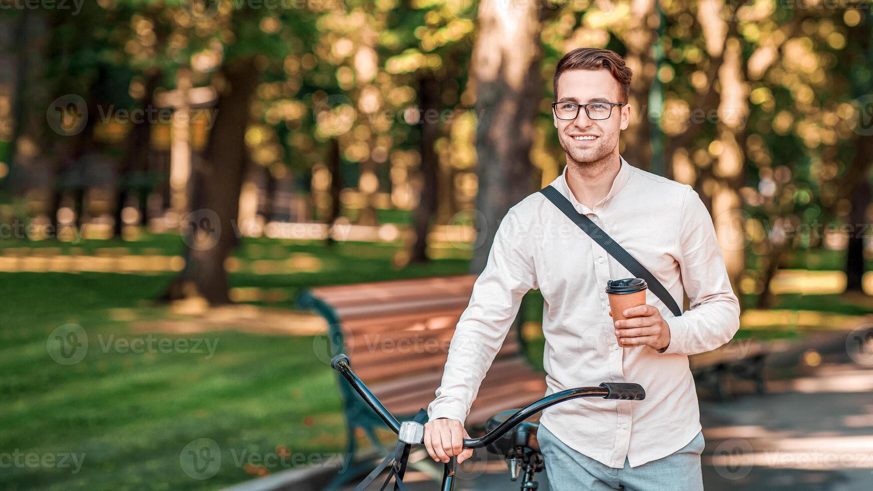 Takeaway coffee and way to work. Attractive happy guy with glasses and cup in hand goes with bike in park, copy space photo