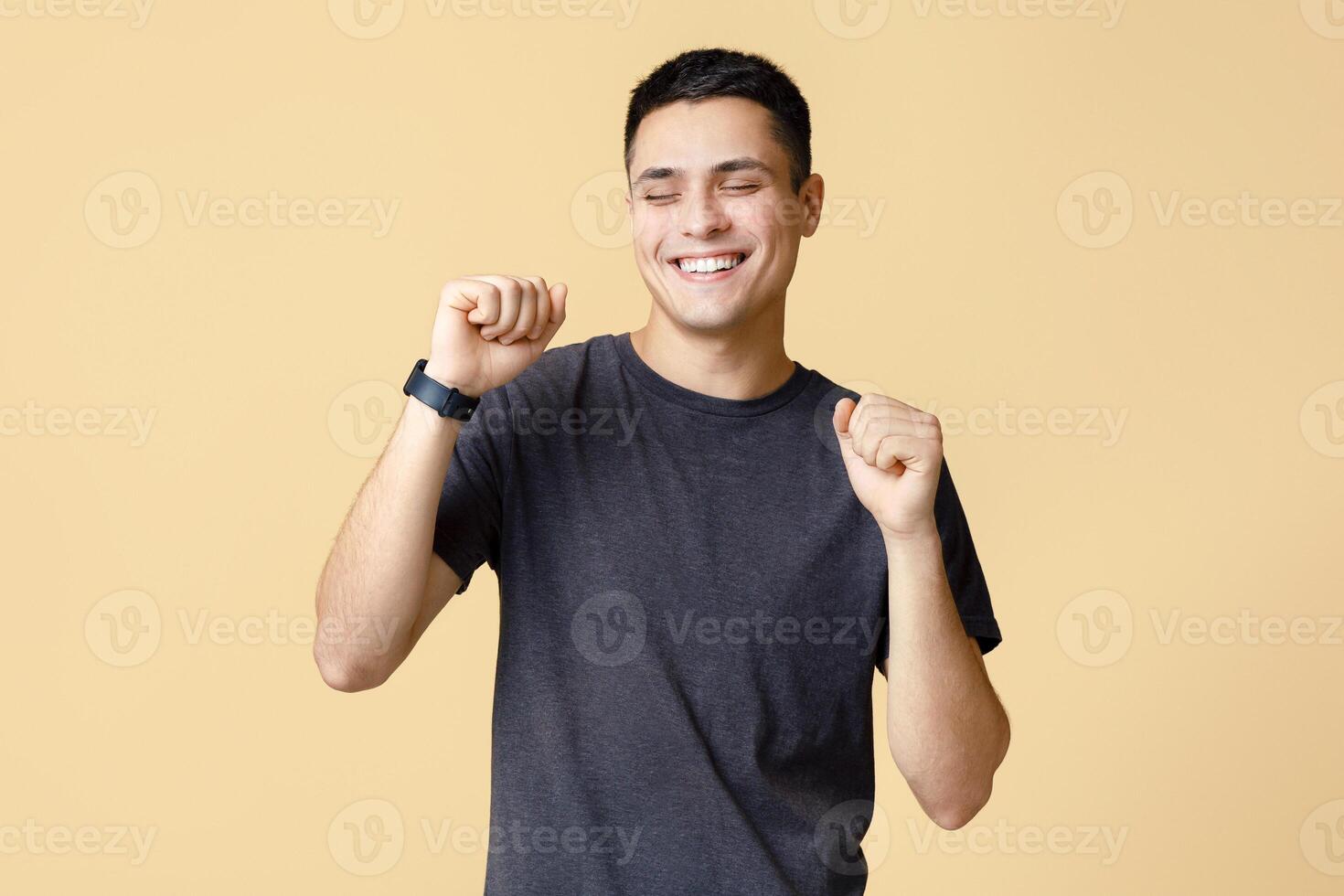 Victory and win dance, positive emotions and great mood. Happy young man student with closed eyes with smart watch expresses success and happiness, isolated on beige background, studio shot photo